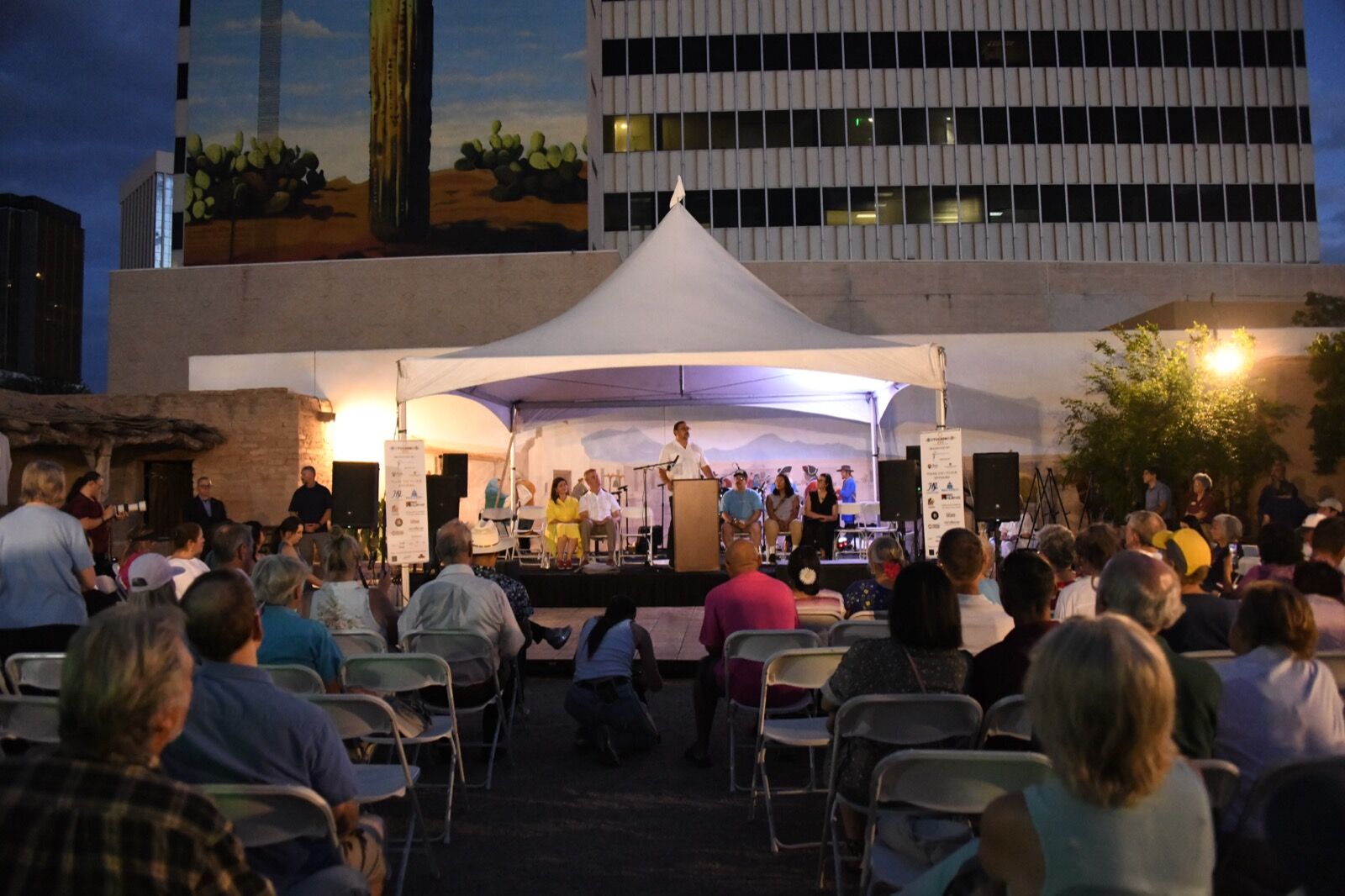 The Presidio plaza at night, a lit white tent stage and a full crowd beneath the saguaro mural and downtown towers