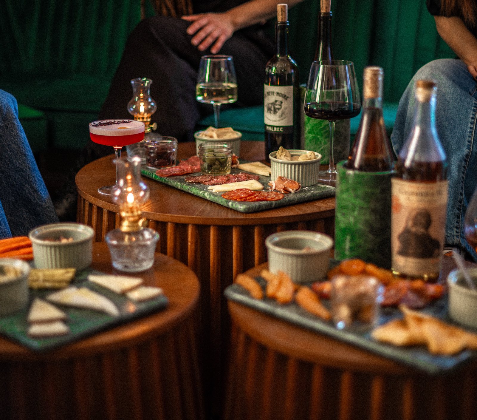 Low tables set with charcuterie, wine and cocktails by oil lamp, guests seated on a green velvet banquette