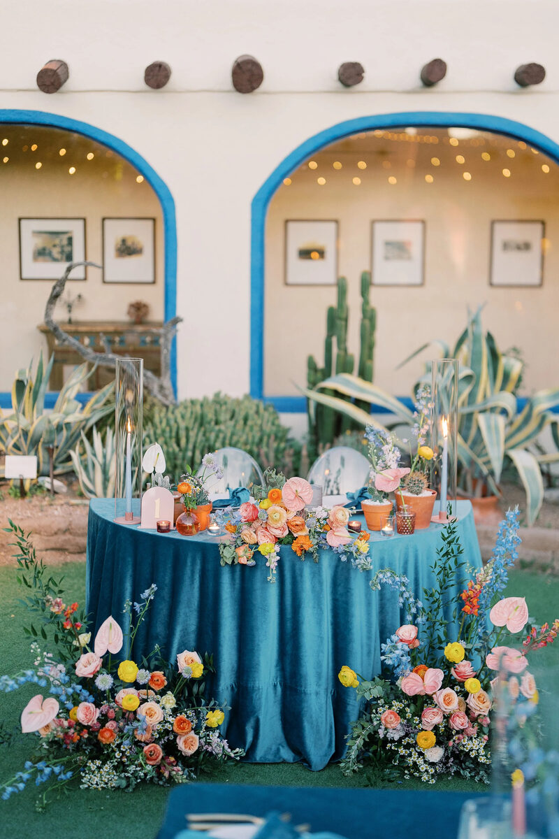 A sweetheart table in teal velvet with bright florals by Bloom Maven, in front of a blue-arched adobe